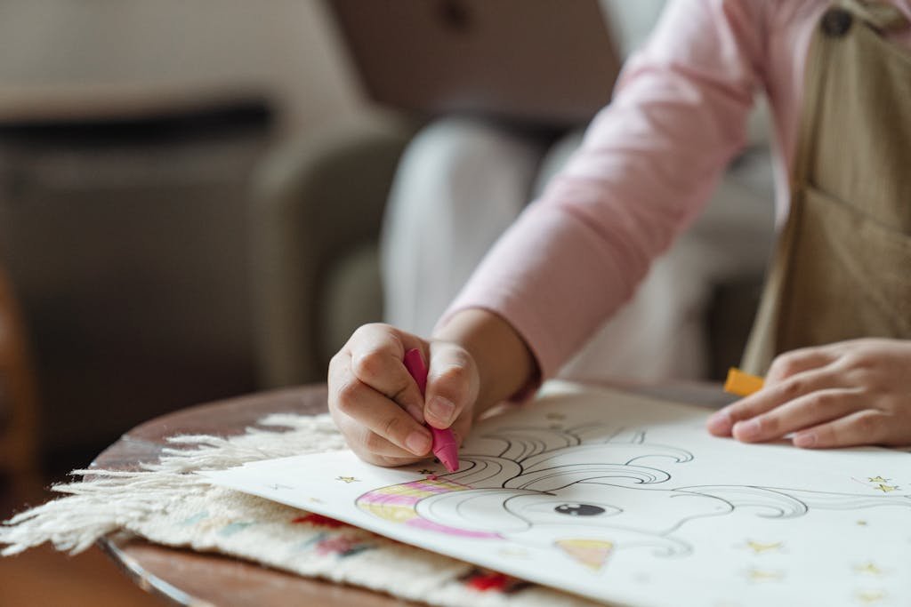 Crop unrecognizable child in light pink turtleneck sitting at coffee table and coloring pictures of coloring book by using multicolored crayons