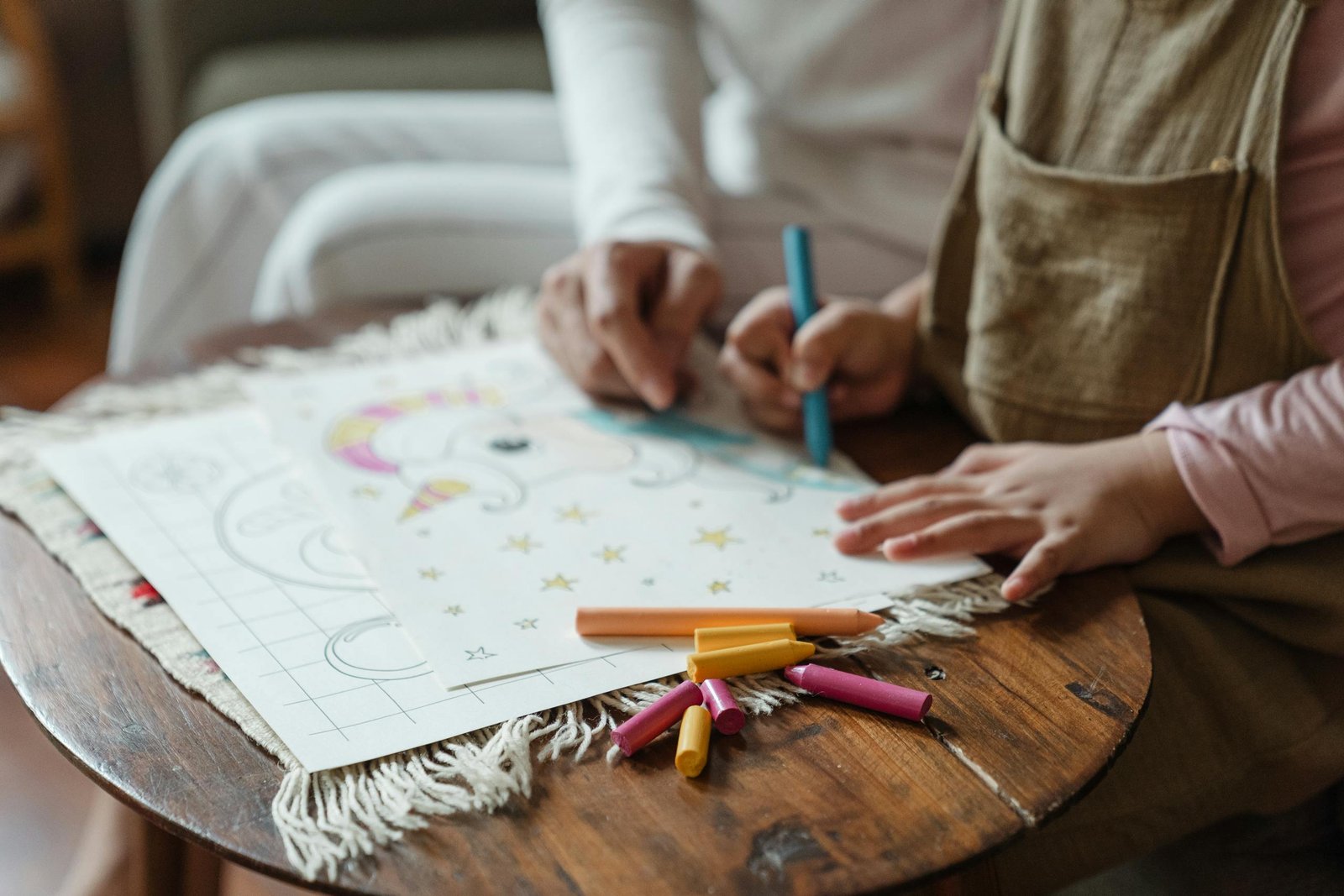 Crop anonymous child and mother sitting at table together and drawing on paper in living room in daylight