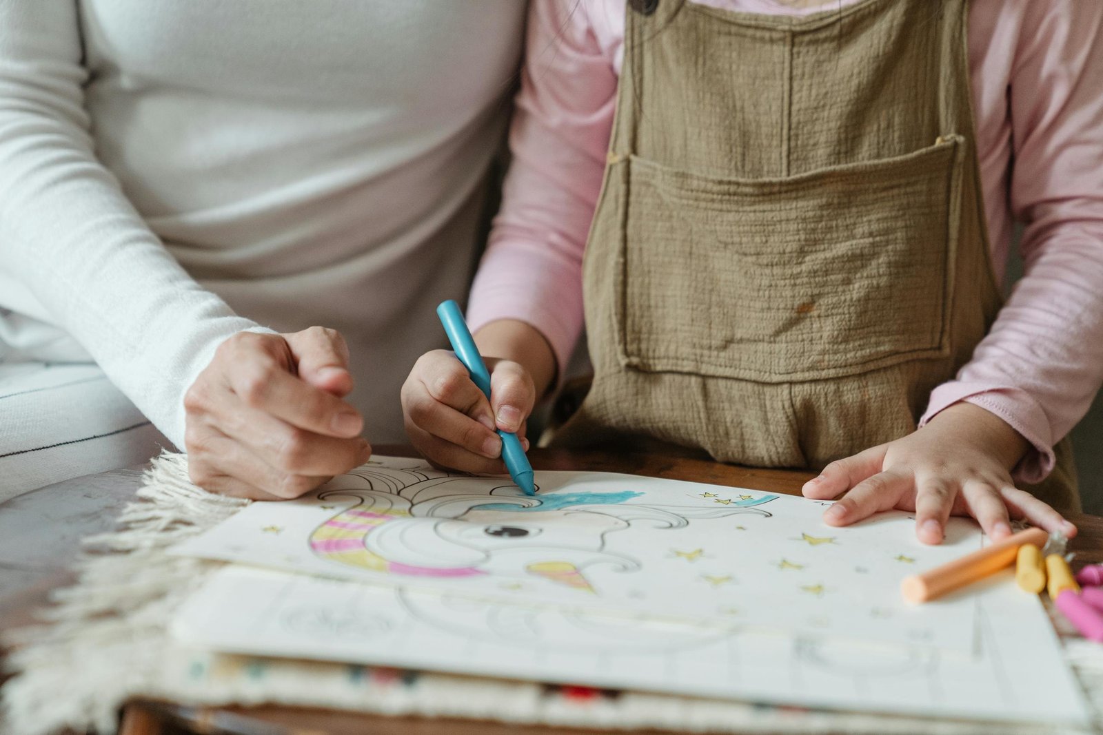Crop anonymous mother with little child in casual clothes sitting at table and drawing with crayons on papers in daylight